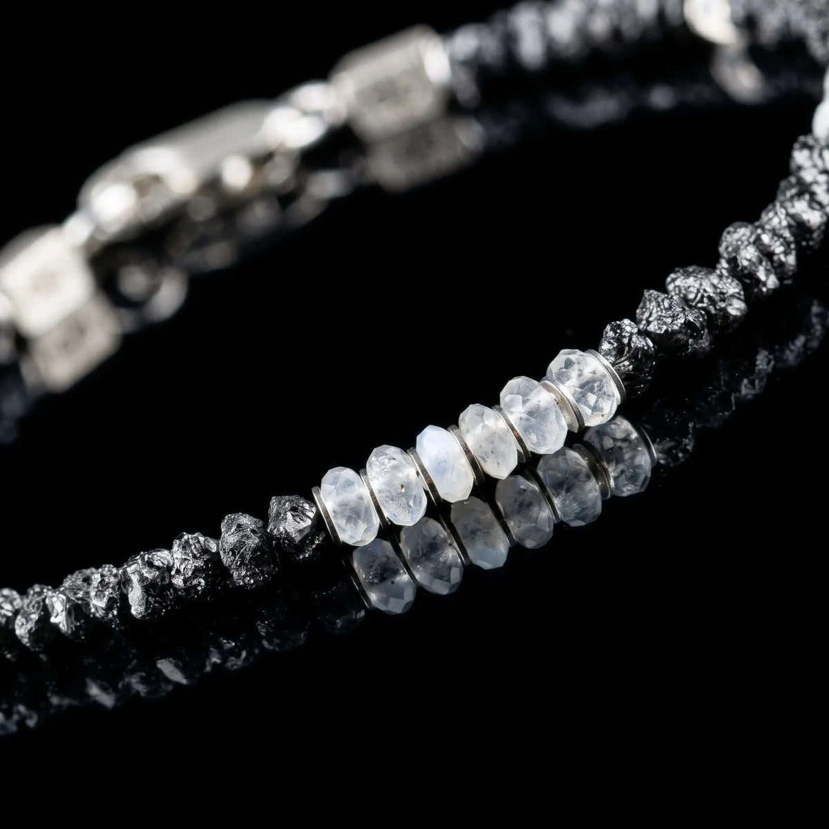 Close-up of Raw Black Diamond - Moonstone Bracelet I (3-4mm), showing pale faceted moonstone between rough black diamond sections on a black reflective surface.