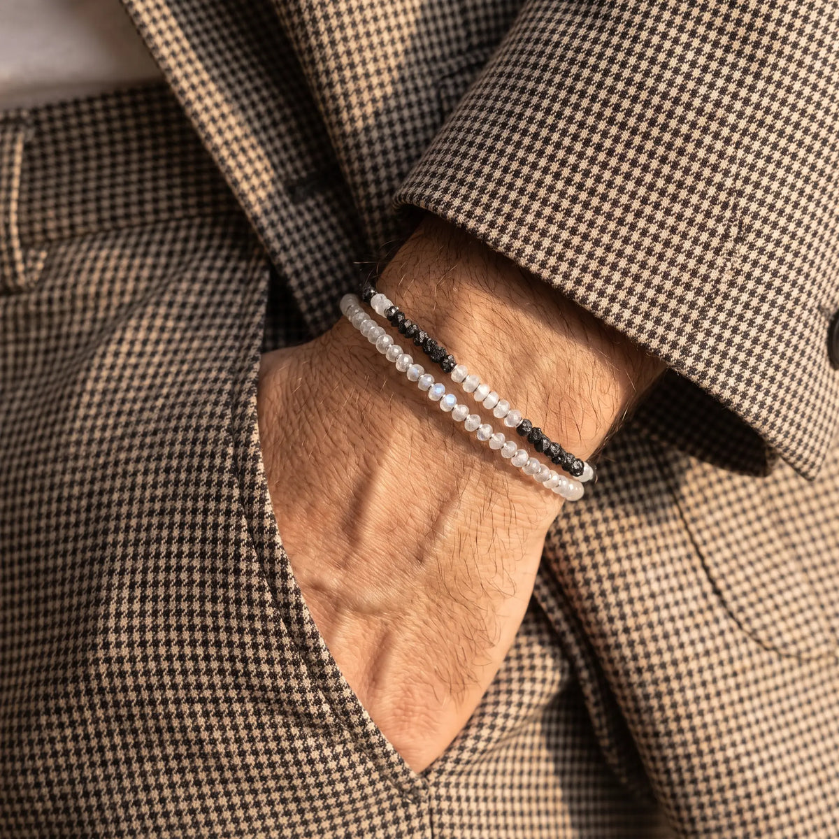 Moonstone Bracelet X (4mm) stacked with Raw Black Diamond - Moonstone Bracelet I (3-4mm) on a wrist beside a brown houndstooth sleeve.